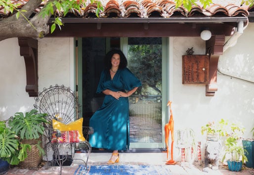 Justina Blakeney stands outside her stucco Spanish Colonial Revival home in front of an open E-Series Hinged Patio Door surrounded by potted plants, a chair, and a giraffe statue.