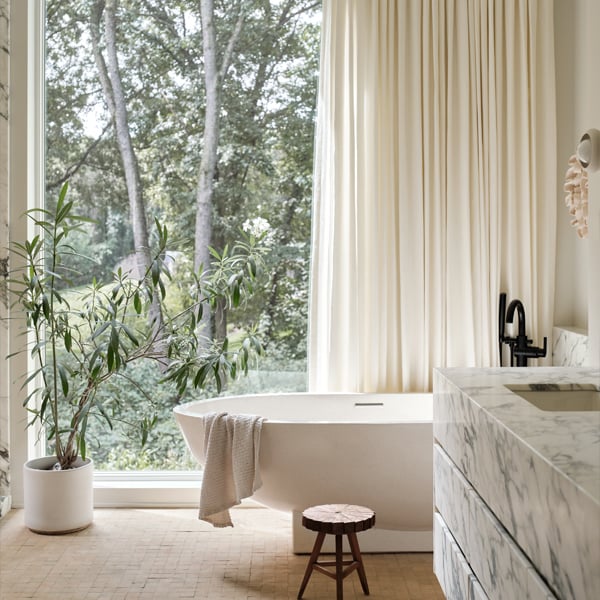 A bathroom with a large soaking tub in front of a floor-to-ceiling window that’s half covered by a white curtain and a floating vanity made of blue-veined marble.